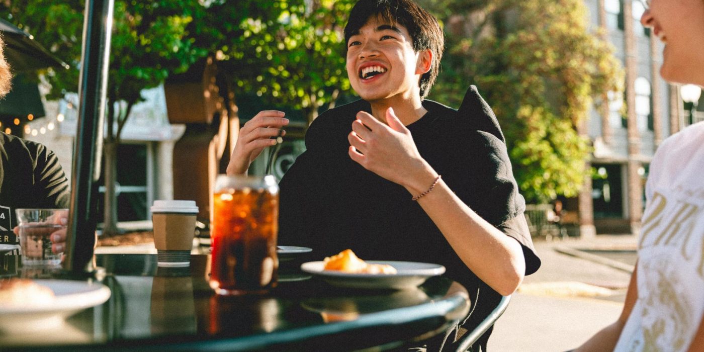 Person smiling and sitting outdoors at a table with iced tea, coffee, and a plate of food, surrounded by greenery.