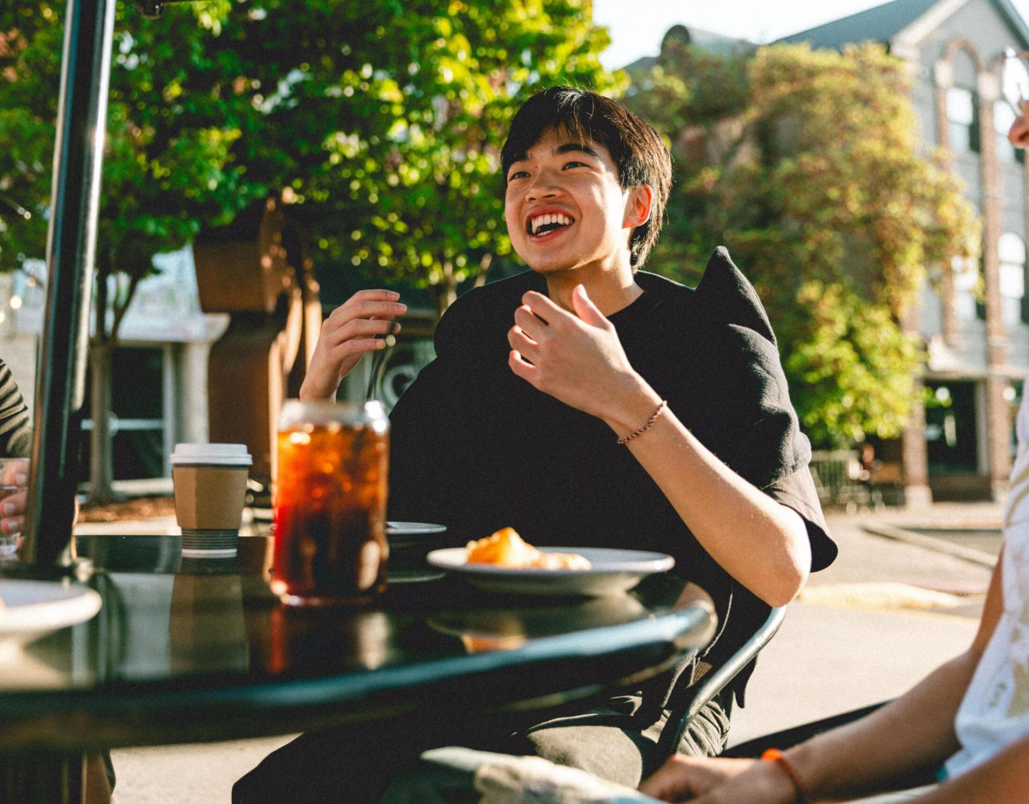 Person smiling and sitting outdoors at a table with iced tea, coffee, and a plate of food, surrounded by greenery.
