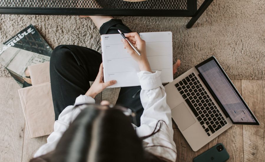 Person sitting and writing into a notebook, applying study tips. They are surrounded by a laptop and books.